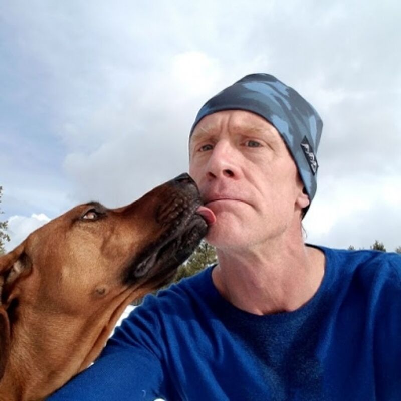 A man in a blue long-sleeved shirt and a camouflage beanie is taking a selfie with his dog. The dog is licking the man's face, and the man has a slightly amused expression. The background shows a cloudy sky and some trees, suggesting they are outdoors. The lighting is bright, indicating it's daytime.